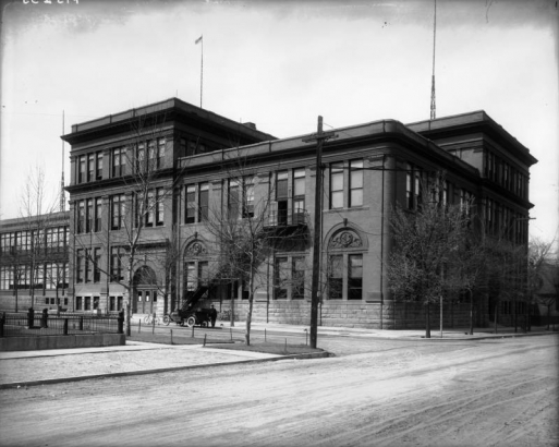A man leans on a car parked in front of Manual Training High School at 27th (Twenty-seventh) and Franklin Streets in the Whittier neighborhood of Denver, Colorado. Shows an Italianate style brick building with tall windows with ornamental arches, dentils, and rusticated first-floor trim.