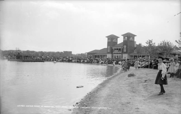 A crowd of people gather around the pavilion and line the shores of City Park Lake (Ferril Lake) in City Park, Denver, Colorado. Men and women are on the balcony of the pavilion. A rowboat and a bandstand are in the lake.