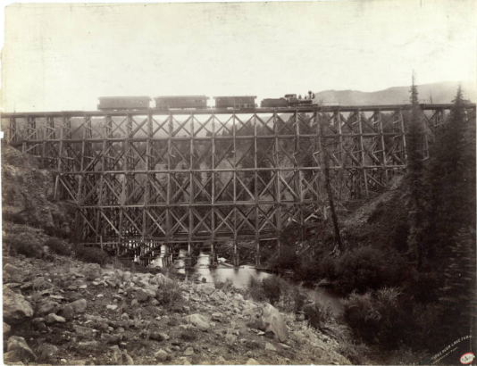 A Denver and Rio Grande narrow gauge locomotive stops on a high wooden trestle over the Lake Fork of the Gunnison River near Lake Fork Junction, Gunnison County, Colorado. Workmen look out from the engine cabin. Shows boulders and brush.