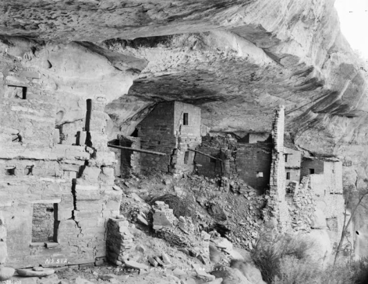 View of Native American (Anasazi) cliff dwelling ruins at Balcony House in Mesa Verde National Park, Colorado. Shows multi-story buildings with windows and stone sills beneath a rock wall. A man, probably a guide, stands near a viga.