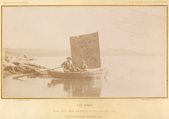 Members of the Hayden Survey (U.S. Geological Survey of the Territories of 1871), sit in a boat, with a tent-fly as a sail and oars, near the shore of Yellowstone Lake, Yellowstone National Park, Wyoming. A canvas drape on the side of the boat reads: "Annie." The boat frame is covered in tar-soaked canvas. Stevenson Island and the Absaroka Range are in the distance.