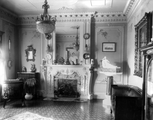 Interior view of the Alonzo Thompson residence (Baerresen Brothers Architects) in Denver, Colorado; the drawing room has French Empire style decor which includes classical entablature, leaded glass, dentils, plaster ornament, a chandelier, a sculpture, a fireplace, torcheres, mirrors, and urns.