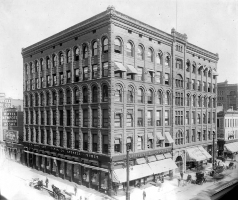 View of the Lewis Mack Building at 16th (Sixteenth) and California Streets in downtown Denver, Colorado. The ashlar stone building has arched windows, storefronts, and signs: "J.S. Appel Suit and Cloak Co.," and "Hotel Newport," and "Morey's Solitaire Coffee." Horse drawn buggies, bicycles, and pedestrians are by the sidewalks.