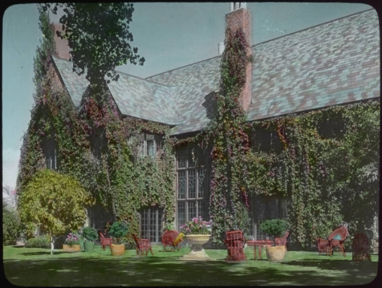 View at the George W. Gano house in Englewood (Arapahoe County), Colorado; landscaping includes a lawn with wicker chairs, urns of flowers, a chestnut tree, and Virginia creeper vines. The house has leaded glass windows and a slate roof.