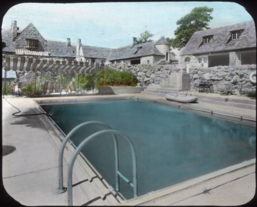 View at Charlford, near Sedalia, Douglas County, Colorado; a manor designed in 1926 for Charles Johnson by Burnham and Merrill Hoyt and built of native volcanic rock. A canvas sun shade and folding chairs are by the swimming pool; the house has a bay window and tower.