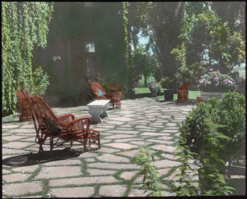 View at the George W. Gano house in Englewood (Arapahoe County), Colorado; landscaping includes flagstone pavement, wicker chairs, a stone bench, and an urn of petunias.