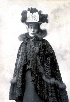 Hispanic American Damiana Rivera de Barela, wife of Colorado State Senator Casimiro Barela, poses for a portrait in an embroidered cape trimmed in fur and a hat decorated with feathers, a bow and a veil.  She wears a pin in the shape of a cross.