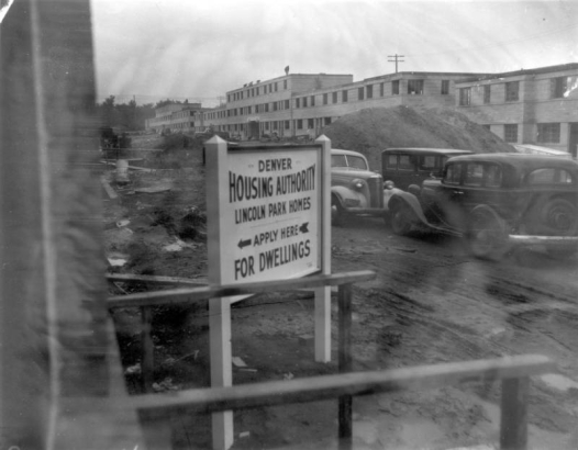 Construction workers roof the Lincoln Park project located  between 13th Avenue and Colfax, and Mariposa and Osage Streets in Denver, Colorado. Shows automobiles and piles of dirt. A sign reads, "Denver Housing Authority, Lincoln Park Homes, Apply Here for Dwellings."