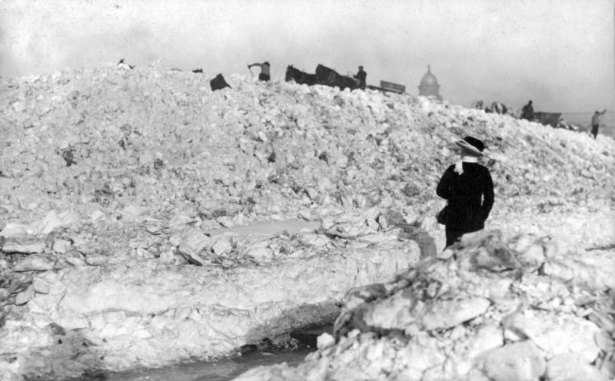 A woman walks near piles of snow dumped by horse-drawn wagons in Civic Center, Denver, Colorado. Shows men at work at the Capitol building in the distance.