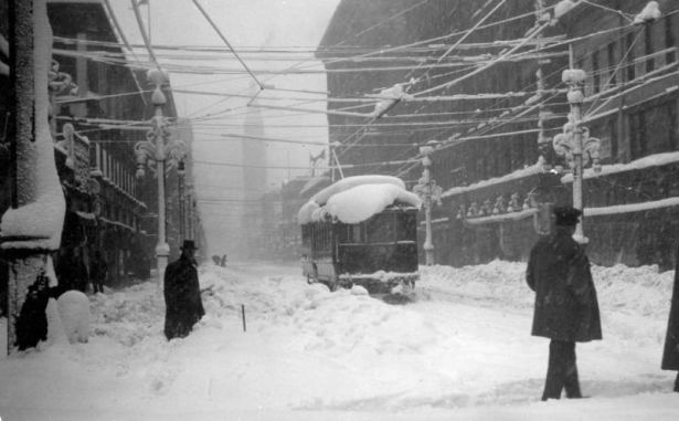 Pedestrians trudge through snow on Welton and Sixteenth (16th) streets in the snowstorm of 1913 in Denver, Colorado. Shows a Denver City Tramway trolley car and snow-laden electrical wires, lamp posts and buildings. The Daniels and Fisher building is in the distance.