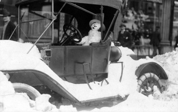 A snowman wears a hat as it sits in the driver's seat of probably a Model T Ford after the snowstorm of 1913 in Denver, Colorado. Pedestrians walk down sidewalk near piles of snow.