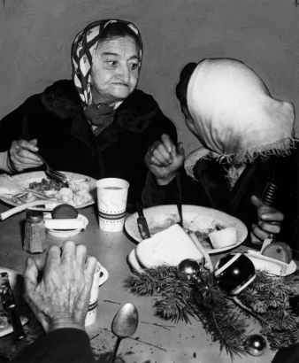 Two women eat Christmas dinner at the Citizen's Mission at 1617 Larimer Street in Denver, Colorado. Shows a Christmas table decoration of pine bough and colored bulbs. The women wear scarves and winter coats.