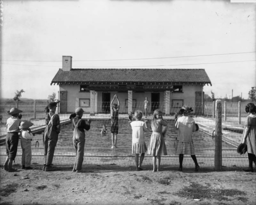 Children stand behind a wire fence and watch a boy in a bathing suit prepare to dive into the Children's Pool at Elyria Playgrounds (Elyria Park) at 48th and High Streets in the Elyria Swansea neighborhood of Denver, Colorado. Shows a stucco building with tile roof and brick quoins. An adult in a swimsuit watches the diver; other swimmers stand in the water and sit at the pool's edge.