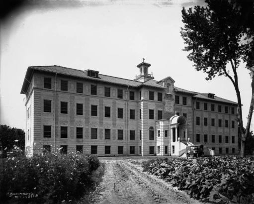 A car sits in front of the dirt driveway of the J.K. Mullen Home for the Aged (later the Little Sisters of the Poor Mullen Home) at 3629 West 29th (Twenty-ninth) Avenue in the West Highland neighborhood of Denver, Colorado. Shows a four story neoclassical building with cupola and portico.