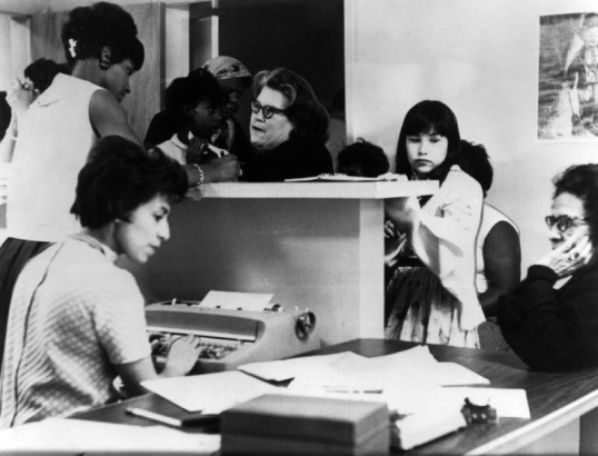 An African American woman helps white and African American clients at the Neighborhood Health Center at 2900 Welton Street in the Five Points neighborhood of Denver, Colorado. Another African American woman types as a woman sits at a table.