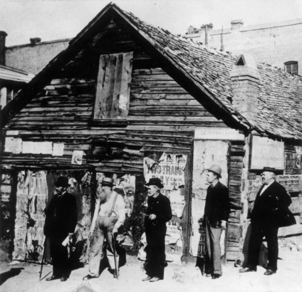Five men stand in front of the Rocky Mountain News building near 1301 Walnut Street in the Auraria neighborhood of Denver, Colorado. Shows a single story wood frame building with brick chimney. Four men wear dark suits and straw boaters or bowlers. One man wears suspenders and leans on a cane. A Midland Railroad advertisment is posted on the side of the building.