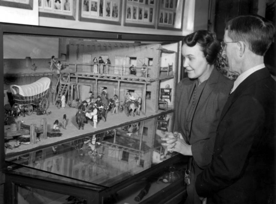 A man and woman examine an exhibit at the Colorado Museum of Natural History in Denver, Colorado. A diorama features a covered wagon, people, horses, and an adobe building with vigas and a balcony.
