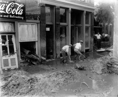 Men use shovels to clear mud from the Cherry Creek flood after the Castlewood Canyon Dam break in Denver, Colorado. Teenage boys look on; storefront signs read: "Coca Cola," "Patterson's Eggs," and "233."