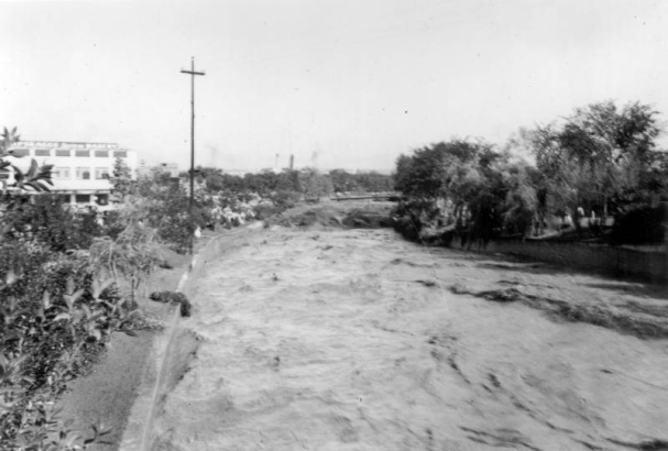 View of a Cherry Creek flood in Denver, Colorado after the Castlewood Canyon Dam break; shows torrents of muddy water. West Denver High School is to the left.