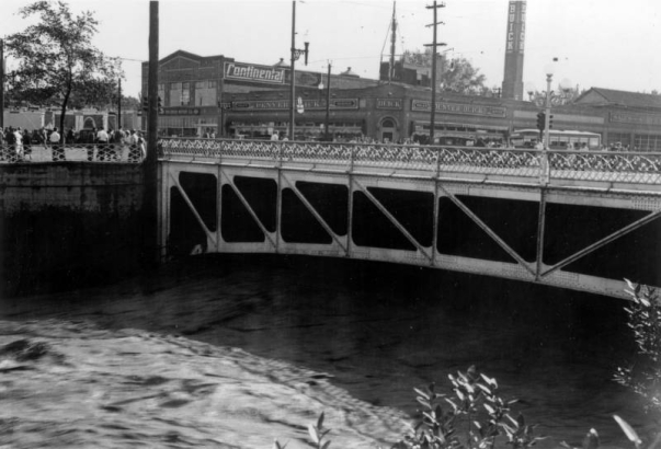 View of a Cherry Creek flood in Denver, Colorado after the Castlewood Canyon Dam break; shows the Broadway Street bridge, people, and a car dealership with a smokestack and signs: "Continental, Buick."