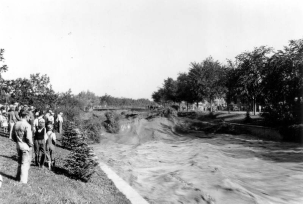 View of a Cherry Creek flood in Denver, Colorado after the Castlewood Canyon Dam break; shows torrents of muddy water in standing waves. People look on from the side.