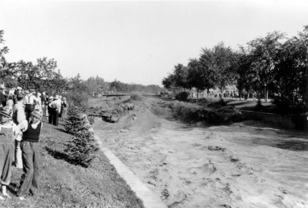 View of a Cherry Creek flood in Denver, Colorado after the Castlewood Canyon Dam break; shows torrents of muddy water in standing waves. People look on from the side; boys wear plastic visors.