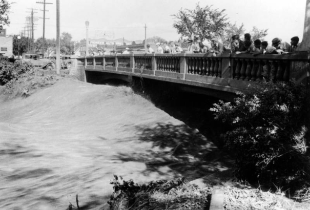 View of a Cherry Creek flood in Denver, Colorado after the Castlewood Canyon Dam break. People stand on the 13th (Thirteenth) Avenue bridge and watch torrents of water.