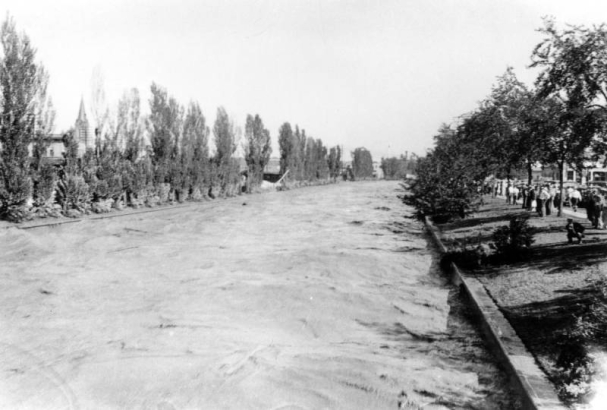 View of a Cherry Creek flood in Denver, Colorado after the Castlewood Canyon Dam break; shows muddy water, landscaping, and people.