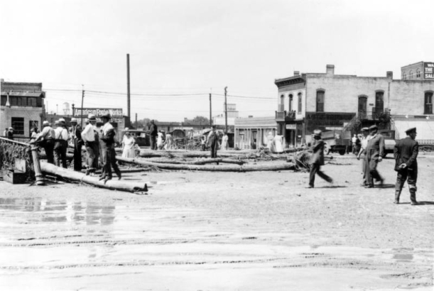 View of a Cherry Creek flood in Denver, Colorado after the Castlewood Canyon Dam break; shows muddy water, the Blake Street bridge, people, debris, and commercial buildings with signs: "American Forge Works."