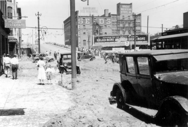 View of Cherry Creek flood damage in Denver, Colorado after the Castlewood Canyon Dam break; shows 16th (Sixteenth) Street, mud, automobiles, storefronts, people, and signs: "Postal Telegraph," "Barteldes Seed Warehouse," "Great Western Stove Co.," and "Salad-Roni."