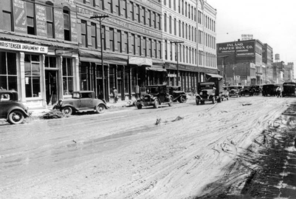 View of Cherry Creek flood damage in Denver, Colorado after the Castlewood Canyon Dam break; shows Wazee Street, mud, automobiles, storefronts, and signs: "Christensen Implement Co," "Oxford Hotel," and "Inland Paper Box Co."