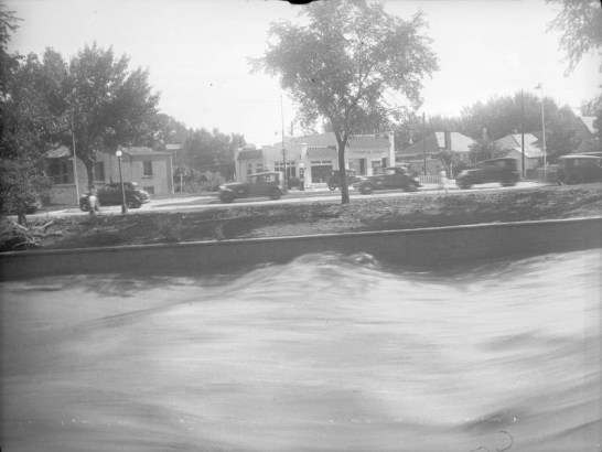 View of the Cherry Creek flood in Denver, Colorado after the Castlewood Canyon Dam break; shows torrents of water, automobiles on Speer Boulevard, and a gas station with tile roof and sign: "Standard Oil Company."