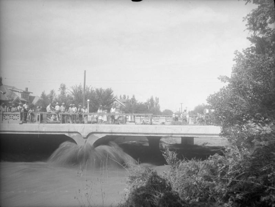 View of the Cherry Creek flood in Denver, Colorado after the Castlewood Canyon Dam break; shows people watching the torrent from a bridge.