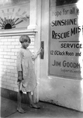 A barefoot girl prepares to open the front door of the Sunshine Rescue Mission at 1822 Larimer Street in Denver, Colorado. The sign in the windows shows a sun with sunbeams that reads: "[Sunshin]e Rescue Mission" and "If [?] Love Shall Conquer Theft." The sign on the door reads: "Hope for all w[?]. Sunshine Rescue Miss[ion]. Service 12 O'clock Noon and [?]. Jim Goodh[eart], Superintende[nt]."