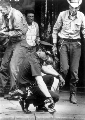 A policeman squats by the side of Larimer Street in the central business district of Denver, Colorado and dumps a bottle of liquor onto the pavement. The police officer wears a short sleeved summer uniform, sun glasses, and leather gloves. A walkie-talkie is in his pocket and keys dangle from his belt. Men on the sidewalk watch the policeman.