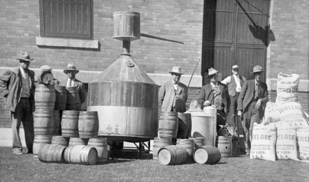 A group of men wearing suits and hats stand near a large still and barrels of liquor near Greeley (Weld County), Colorado. One man leans his arm on a pile of sacks with labels reading: "100 lbs, Cerelose, Product Refining Co., New York, U.S.A."