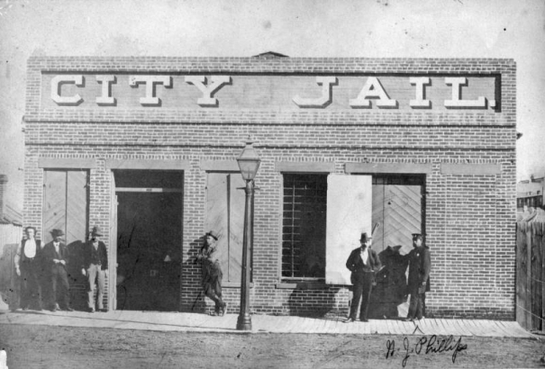 A group of men stand outside the Denver City Jail located at 231 Thirteenth (13th) Street, Denver, Colorado. A police officer stands outside a set of large wooden doors.  The building is a one story brick structure with heavy wood shutters. Bars cover the windows. Large letters painted on the front of the building read: "City Jail."