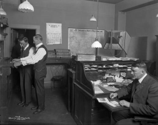 A clerk presses the inked finger of a man onto a paper at the Denver Police Department offices in the City Hall Building, located at Fourteenth (14th) Street and Larimer Street in Denver, Colorado. Another man sits at a roll top desk near a photographic lamp and backdrop.  A map labeled "Chicago & North Western Line" hangs on the wall. A calendar advertising "The Continental Life Insurance Co." turned to March 1929 hangs nearby. Light fixtures hang from the ceiling.