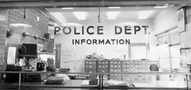 Police officers sit at desks at the information office in the Denver Police Building, Denver, Colorado. A glass shield labeled "Police Dept. Information," runs along the front of the desk. Shows file cabinets, telephones and typewriters.