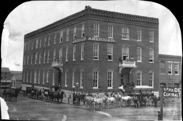 People, horses, wagons, and buggies stand outside the American House, a hotel at 16th (Sixteenth) and Blake Streets in Denver, Colorado; other people stand on balconies and on the roof. The hotel is a three-story brick lodging house with stone trim, a covered entry, and adjacent storefronts. A sign on the hotel reads: "Charlie North's Billiard Rooms."