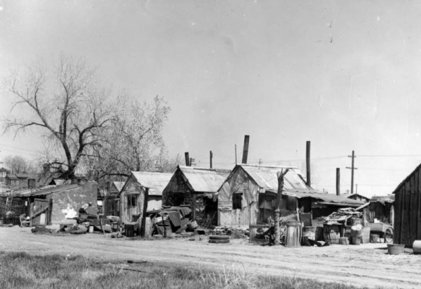 View of makeshift houses and shanties at 19th (Nineteenth) and Clay Streets in the Jefferson Park neighborhood of Denver, Colorado. A man works by barrels and corrugated metal.