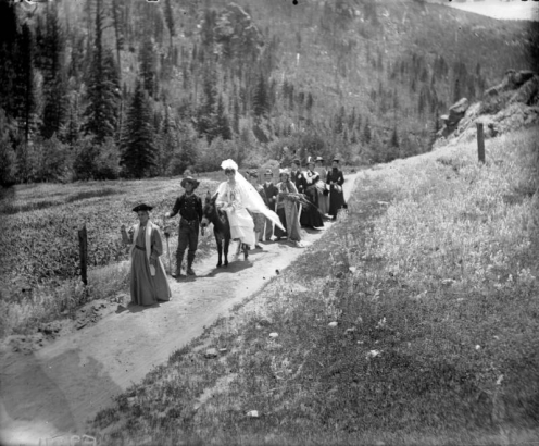 View of a wedding procession in the Colorado mountains; the bride wears a horseshoe, veil, and gown and rides a burro. The groom wears a Civil War uniform; well-dressed people follow.