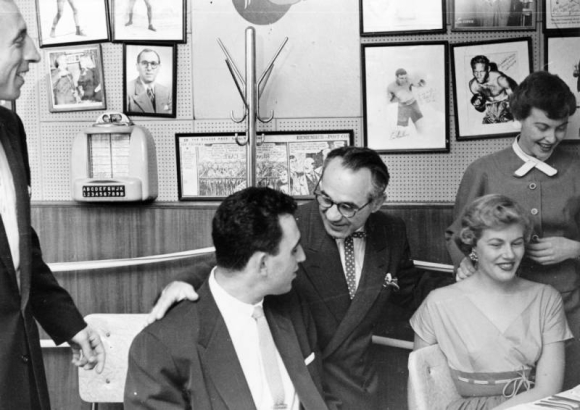 Interior view at the Ringside Lounge in Denver, Colorado; shows Joe "Awful" Coffee (with glasses), members of the Denver Bears baseball team, and women. Photographs of boxers, a comic strip, and a juke box selector are on the wall.