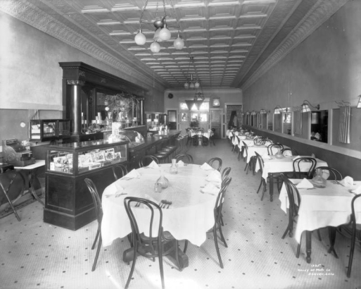 Interior view of the City Cafe restaurant in Denver, Colorado; decor includes a carved wood bar, a tile floor, potted plants, lamps, coving, United States flags, a glass display case of cigars, and a pressed tin ceiling.