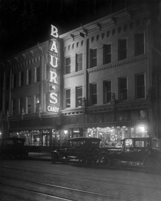 Night view of 1512 Curtis Street businesses in downtown Denver, Colorado; electric sign reads: "Baur's Candy." Cars are by the storefront windows.
