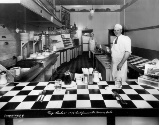 Interior view of the Pig Parlor restaurant in downtown Denver, Colorado; men pose in the kitchen behind a checkered counter and condiments. Decor includes a deco ceiling lamp, a mural of pigs, a grill, coffee machines, and a menu.