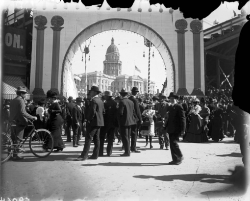People walk in a baseball stadium (probably Broadway Athletic Park) at Colfax Avenue and Broadway in Denver, Colorado, during the Festival of Mountain and Plain. The Colorado State Capitol building shows through the entry arch.