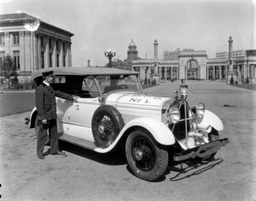 Fire Chief John F. Healy poses in uniform by a Fire Department vehicle (Lincoln sedan) in Civic Center park, Denver, Colorado. The automobile has a bell and siren. The Voorhies Memorial Colonnade, Carnegie Library, and Arapahoe County Courthouse dome are in the background.