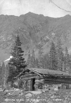 A miner, in a striped suit with a pick axe, poses next to a log cabin in possibly Clear Creek County, Colorado. The cabin has a gabled roof, a metal chimney, a dirt roof and a sign in a window that reads: "[C]rocker [& Co.] Crac[kers], Denver." Trees are near the cabin, steep slopes and talus are on mountains above timberline.