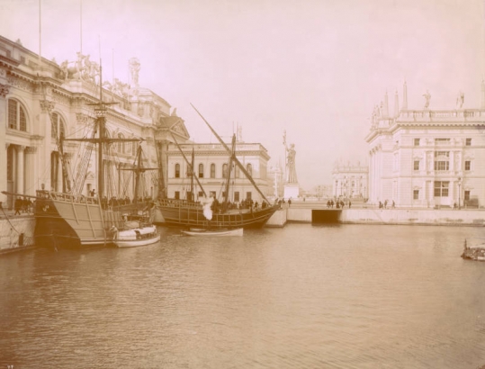 Reproductions of Columbus' ships (caravels) the Nina and the Pinta are moored in the South Pond at the World's Fair (World's Columbian Exposition) in Chicago, Illinois.  Spectators stand on the sidewalk near the Agricultural Building. The Statue of the Republic is in the distance. Print 88.